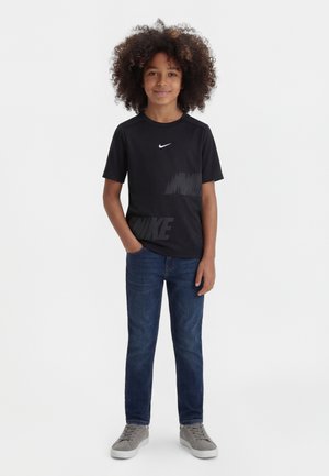 Băiat cu păr creț, purtând un tricou negru Nike, blugi albaștri și adidași gri, stând cu o mână în buzunar, pe un fundal alb.