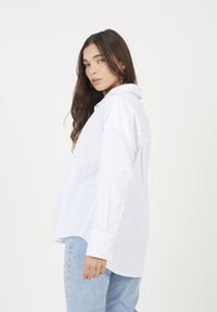 Donna con lunghi capelli castani che indossa una camicia bianca sbottonata e jeans azzurri chiari, in piedi di profilo contro uno sfondo bianco semplice.