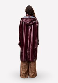 Lange bordeauxrode waterdichte regenjas met een capuchon, met een glanzende textuur en een contrasterend grijze binnenkant. Gecombineerd met brede geruite broeken.