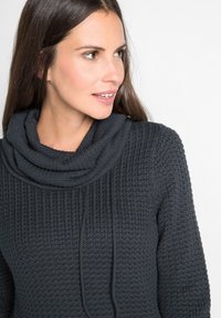 Maglione lavorato a maglia grigio scuro con collo a scatola, tessuto testurizzato e dettaglio con cordoncino. Primo piano della zona del collo e della spalla visibili.