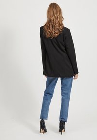 Blazer noir sur mesure à coupe droite, porté avec un jean bleu clair. Il possède des manches longues et est associé à des bottes noires à talons hauts.