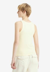 Geribbelde gele tanktop met een strakke pasvorm en brede schouders. De achterkant heeft een ronde halslijn en een gladde textuur. Gecombineerd met crèmekleurige shorts.