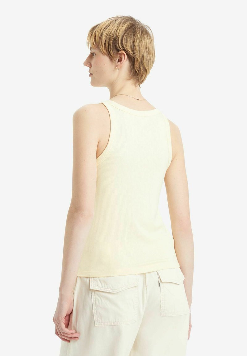 Rippenshirt in Gelb mit tailliertem Design und breiten Trägern. Der Rücken hat einen runden Ausschnitt und eine glatte Textur. Kombiniert mit cremefarbenen Shorts.