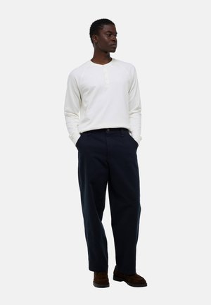 Homme debout, mains dans les poches, portant une chemise henley blanche à manches longues, un pantalon bleu marine et des chaussures marron, légèrement tourné vers la gauche.