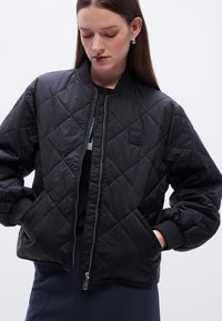 Μαύρο μπουφάν bomber με καπιτονέ σχέδιο, φερμουάρ μπροστά, ριμπ μανσέτες και λογότυπο. Διαθέτει ομοιόμορφη, γυαλιστερή υφή και άνετη εφαρμογή.