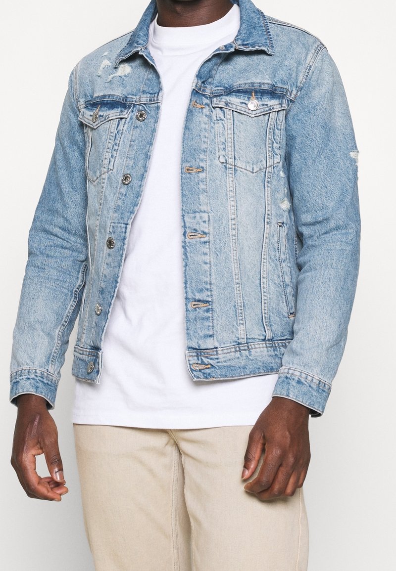 Světle modrá džínová bunda s klasickým designem, vybavená dvěma náprsními kapsami, opotřebovanými detaily a kovovými knoflíky. Nositelná přes bílé tričko.