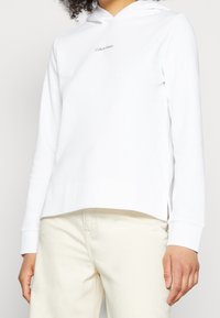 Sudadera blanca de algodón con bolsillo tipo canguro, mangas largas y capucha con cordón. Sutil logo de "Calvin Klein" en el pecho.