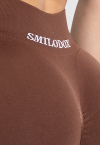 Bruna högmidjade leggings tillverkade av ribbat material, med en vit "SMILODON"-logotyp längs midjebandet och en mjuk yta.