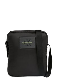 Bolso de hombro rectangular negro hecho de nailon, con un parche frontal texturizado que lleva el logotipo "CK" en relieve, correa ajustable y cierre de cremallera.