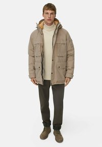 Beige gepolsterte Jacke mit Kapuze, zwei Brusttaschen und einem kontrastierenden orangefarbenen Innenfutter, getragen über einem hellen Pullover und dunklen Hosen.