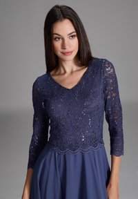 Marine blaues Kleid mit einem Spitzenoberteil und Paillettenverzierungen, V-Ausschnitt und Dreiviertelärmeln, das in einen weichen Stoffrock übergeht.