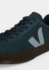 Baskets en daim bleu foncé avec lacets foncés, semelle en caoutchouc marron et grand logo "V" gris clair sur le côté.