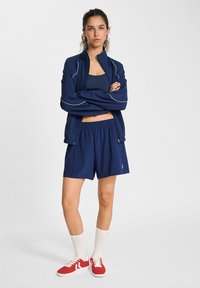 Giacca sportiva navy con accenti bianchi, abbinata a pantaloni corti coordinati. Il modello indossa scarpe da ginnastica rosse e calze bianche fino al ginocchio.