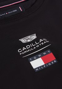 Μαύρο ύφασμα με λευκό λογότυπο της ομάδας Cadillac Formula 1 πάνω από το λογότυπο της σημαίας Tommy Hilfiger σε κόκκινο, λευκό και μπλε.