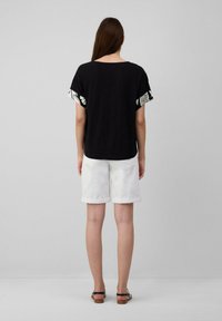 Schwarzes T-Shirt mit gemusterten kurzen Ärmeln, kombiniert mit weißen Shorts mit umgeschlagenem Saum. Das Outfit wird mit einfachen Sandalen abgerundet.
