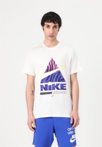 Balta kokvilnas t-kreklu ar purpura un rozā trīsstūra logotipu, kurā ir zebru svītras un teksts "NIKE", saskaņots ar zilām šortiem.