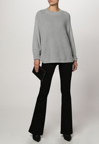 Šedý pletený svetr s klasickým výstřihem a žebrovanou strukturou, doplněný černými rozšířenými kalhotami a elegantními černými botami, držící černou kabelku.