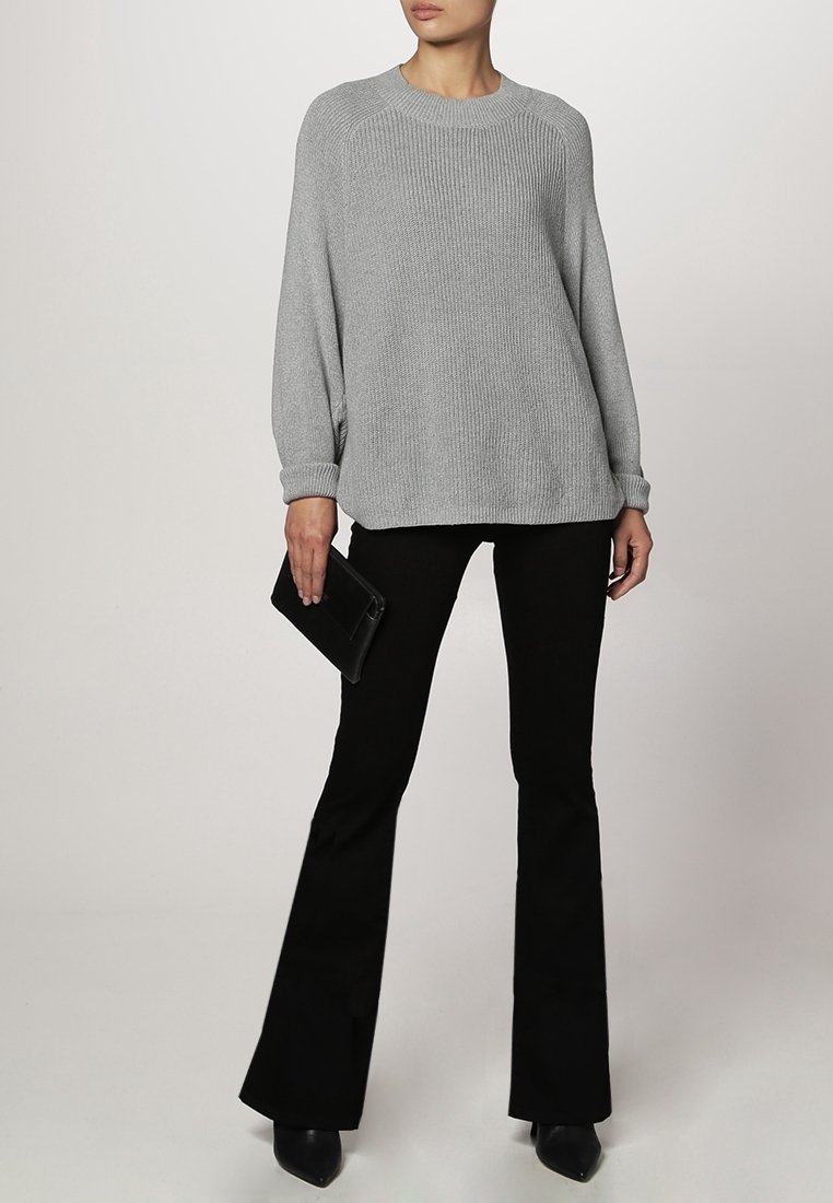 Šedý pletený svetr s klasickým výstřihem a žebrovanou strukturou, doplněný černými rozšířenými kalhotami a elegantními černými botami, držící černou kabelku.