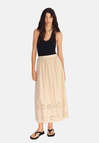 Maxi gonna color beige chiaro con motivi circolari ricamati all'orlo, vita arricciata e una silhouette fluida, abbinata a un top nero e sandali.