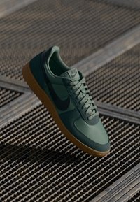 Grønn Nike-sneaker med teksturerte materialer, svart swoosh og beige yttersåle. Har runde lisser og polstret krage.