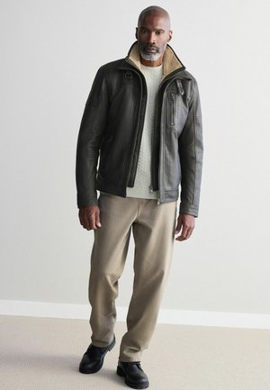 Homme portant une veste en cuir noire avec un col en laine, un pull crème à torsades, un pantalon beige et des chaussures noires, debout contre un mur uni.