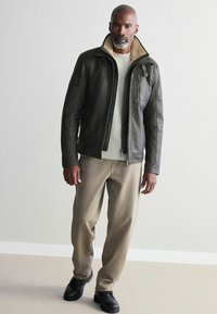 Chaqueta de cuero marrón con cuello alto y forro de felpa crema, combinada con un suéter ligero, pantalones color beige y zapatos negros.