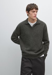 Jonge man in een donkergrijze gebreide trui over een wit shirt en zwarte broek, staand met zijn handen op zijn rug tegen een effen achtergrond.