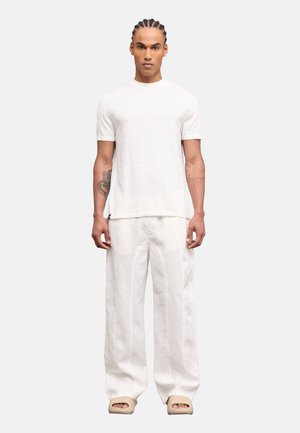 Hombre con cabello trenzado que lleva una camisa blanca de manga corta, pantalones blancos sueltos y sandalias beige abiertas en los dedos, de pie derecho.