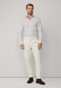 Weißes gemustertes Hemd mit hellblauen Akzenten, kombiniert mit hellbeigen Hosen und dunkelbraunen Stiefeletten. Schlicht, taillierte Passform.
