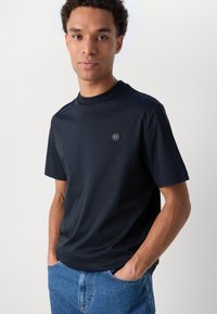 T-shirt à col rond bleu marine en tissu doux, arborant un petit logo en relief sur la poitrine, associé à un jean bleu clair.