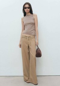 Brown fitted sleeveless top paired with wide-leg beige pants featuring a chain belt and a small brown handbag. Smooth textures throughout.