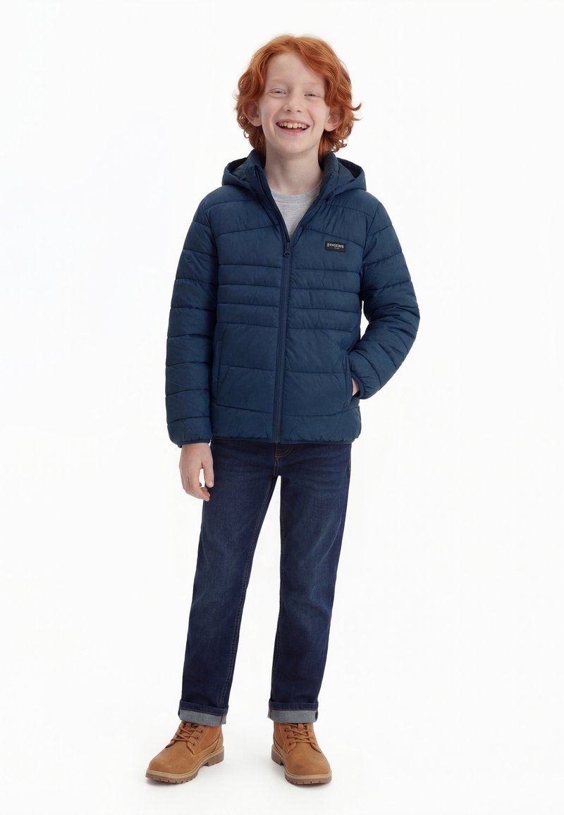 Garçon aux cheveux roux souriant, portant une veste matelassée bleu marine, une chemise grise, un jean foncé et des bottes à lacets couleur beige, debout avec une main dans la poche.