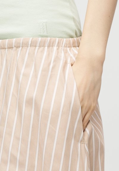 Main reposant dans la poche d'un pantalon beige et blanc à rayures verticales avec une taille élastique et un haut vert clair.