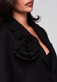 Černý blazer s texturovanou látkou, s velkým černým látkovým růžovým akcentem na klopě. Klasický design s výraznými švy.