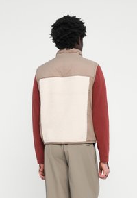 Padded vest with a light brown outer layer and white fleece back panel, featuring a high collar and zip closures.
