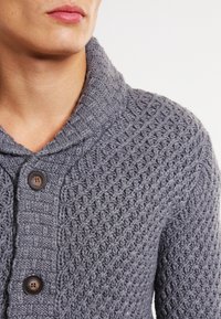 Detailní záběr osoby nosící texturovaný šedý pletený kardigan se dvěma tmavými knoflíky a šálovým límcem.