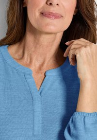 Eine Frau trägt ein blaues Strickshirt mit einem tiefen Ausschnitt, legt ihre Hand an ihr Gesicht und lächelt leicht mit glänzenden Lippen.