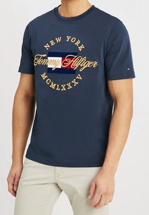 Homme portant un t-shirt bleu marine Tommy Hilfiger avec le texte New York et un logo rouge, blanc et bleu, associé à un pantalon beige clair.