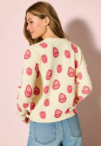 Cremefarbener Pullover mit rosafarbenen Kuchenmustern, rundem Ausschnitt und gerippten Bündchen. Kombiniert mit hellblauen Denim-Jeans.