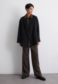 Zwart dubbelzijdig jasje, een bruin shirt met stippen en een brede broek met stippen, gecombineerd met zwarte platform schoenen op een neutrale achtergrond.
