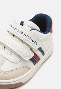 White leather sneaker featuring navy and burgundy stripes, Velcro straps, rounded toe, and a textured rubber sole. Brand name visible.