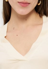 Χείλη, λαιμός και πάνω μέρος του στήθους ενός ατόμου που φορά χρυσούς κρίκους στα αυτιά και κορυφή με V λαιμόκοψη σε εκρού χρώμα, με εμφανείς ελιές στο δέρμα.