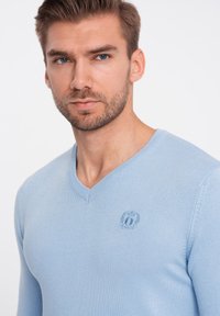 Hellblaues V-Ausschnitt-Pullover aus Strickstoff. Verfügt über ein dezentes gesticktes Logo auf der Brust und Rippdetails am Kragen und an den Ärmeln.
