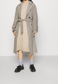 Pasiasty długi płaszcz z paskiem, beżowa dzianinowa sukienka pod spodem, lawendowe skarpetki oraz czarne buty z grubą podeszwą i akcentem w postaci kokardki.