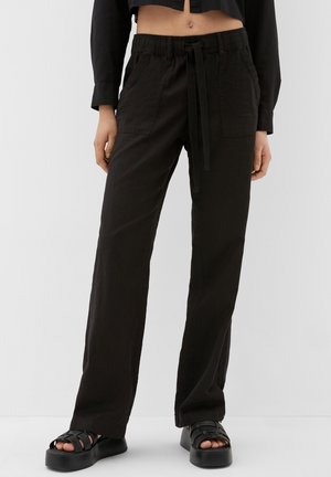 Persona con pantalones negros de pierna ancha con cintura de cordón y sandalias negras de plataforma, de pie frente a un fondo claro y liso.