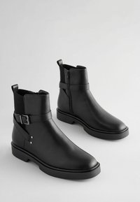 Bottines en cuir noir avec des panneaux élastiques sur les côtés, détail de boucle et semelles plates. Texture lisse, bout arrondi et fermeture éclair discrète.