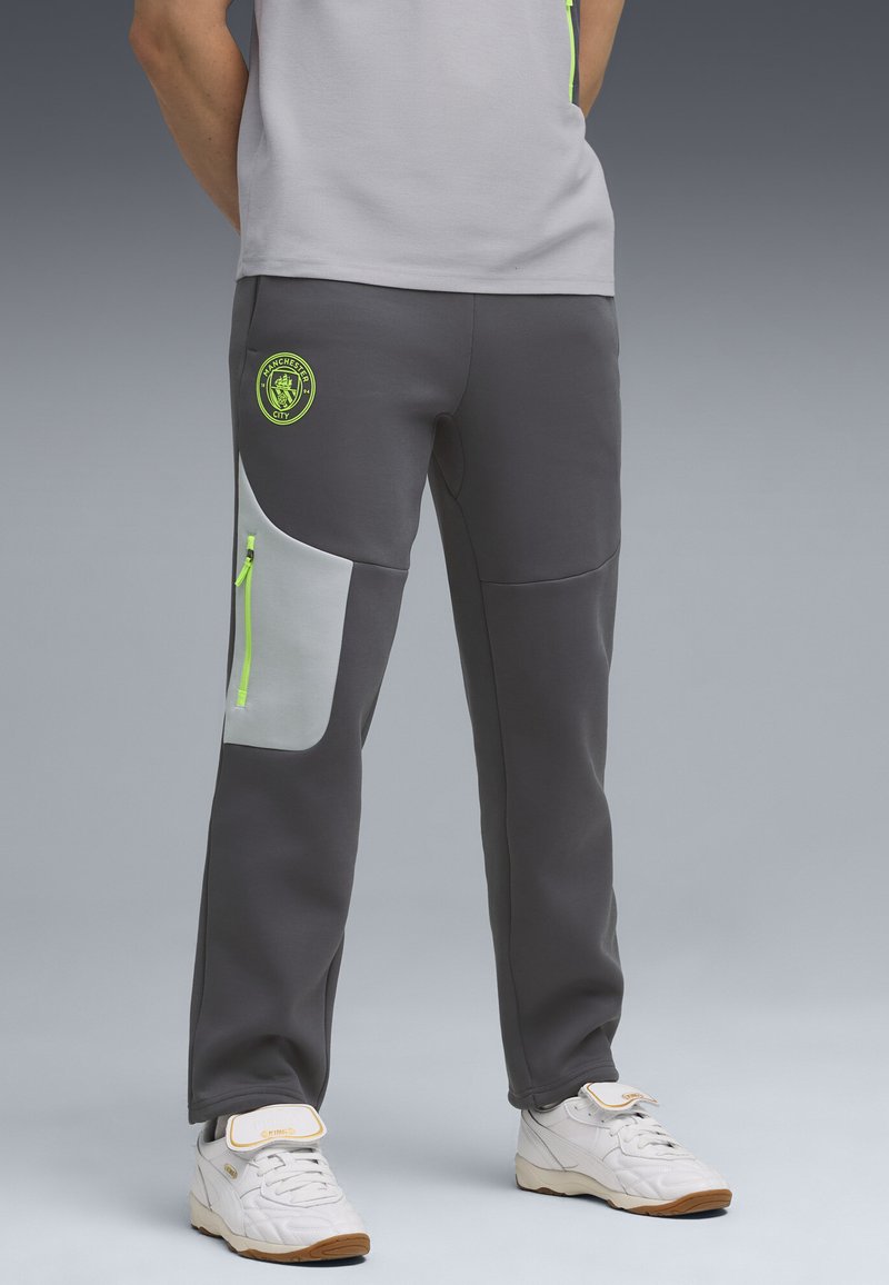 Grijze sportieve joggers met een lichtgrijze zijpaneel, neon groene accenten, elastische tailleband en een Manchester City-logo op de bovenbeen.