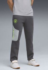 Šedé športové nohavice so svetlošedým bočným panelom, neonovými zelenými akcentami, elastickým pásom a logom Manchester City na stehne.