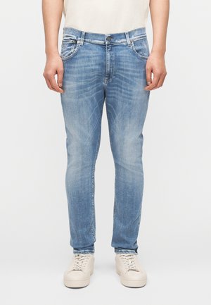 Asmuo, dėvintis šviesiai mėlynas siauras džinsus ir smėlio spalvos sportinius batelius, stovintis priešais paprastą baltą foną.