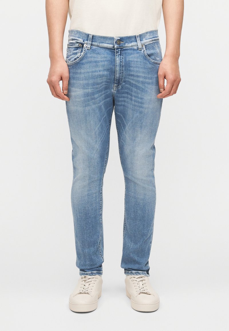 Άτομο που φοράει ανοιχτό μπλε τζιν στενής εφαρμογής και μπεζ αθλητικά παπούτσια, στέκεται μπροστά σε λευκό φόντο.